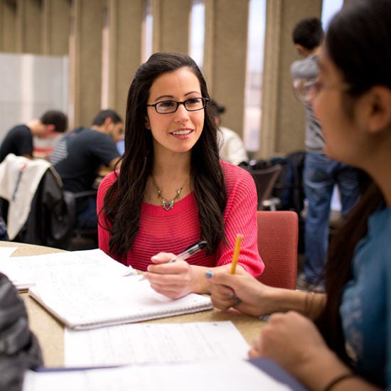 Two students sit at a table working together. They have notebooks and pens. There are other students in the background also studying.