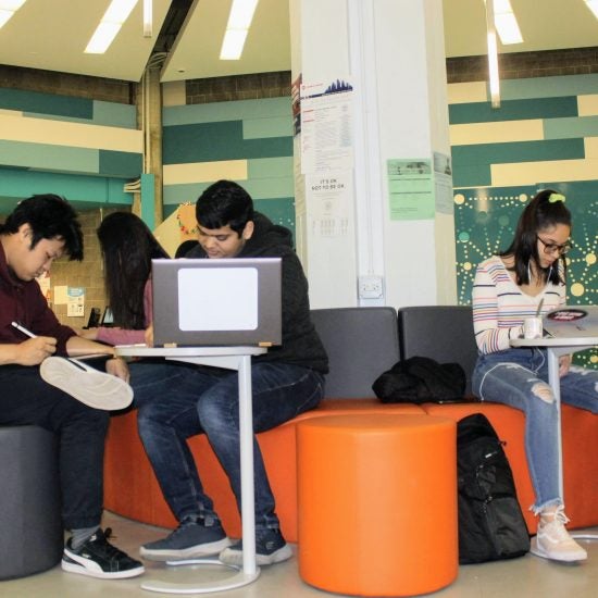 A few students sit on the gray and orange benches and ottomans surrounding the column in the center of the MSLC. There are small moveable tables that they are working on.