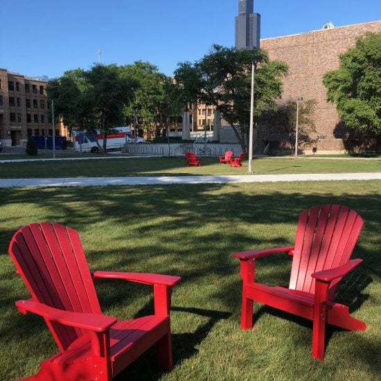 Two bright red lawn chairs are angled toward each other on the green lawn of UIC's campus. In the background there are buildings, trees, and a clear blue sky.