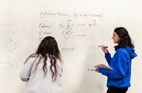 Two students stand facing a whiteboard wall in the MSLC. One student has their back turned to the camera and appears to be writing on the whiteboard. The other is turned so their side profile is visible and is holding two markers and a tablet. There is writing on the whiteboard that says 