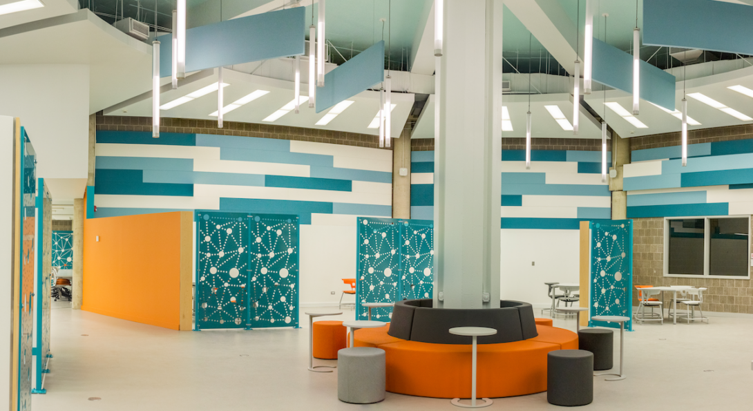 View of center seating area of MSLC. There is a column in the center, surrounded by gray and orange seating and small moveable tables. Behind it, there are different sections with tables and chairs.