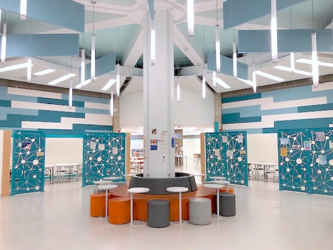 View of center seating area of MSLC. There is a column in the center, surrounded by grey and orange seating and small moveable tables. Behind it, there are different sections with tables and chairs.